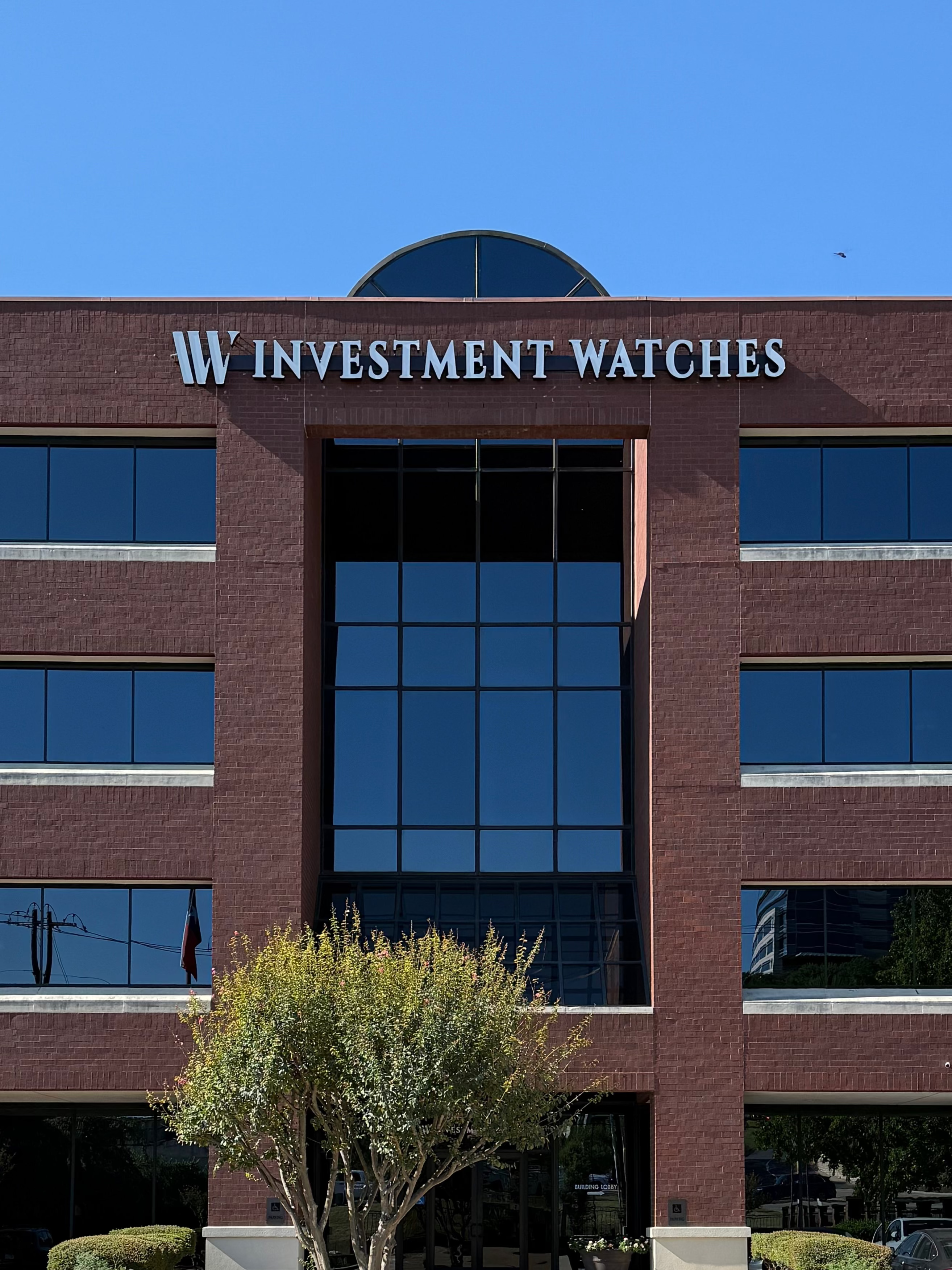Brick office building with 'Investment Watches' sign, American flag, and parking lot.